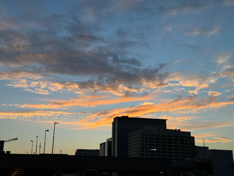 Orange clouds of sunset and the silhouette of a large building and unlit streetlights.